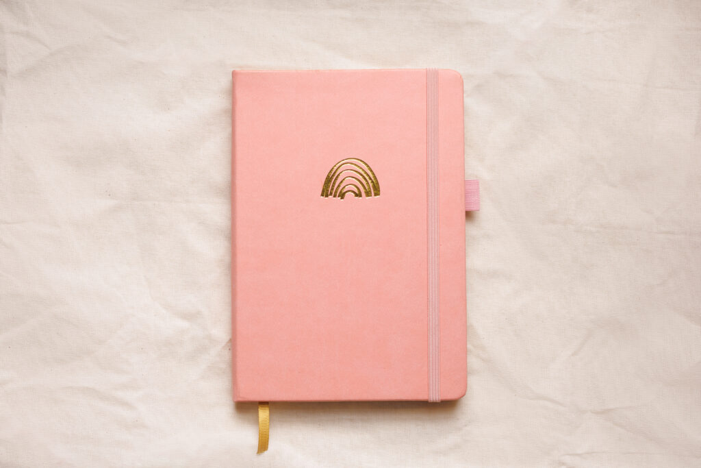 Caderno A4 com capa rosa, fitilho dourado e um foil dourado em formato de arco-íris na capa, usado para bullet journal do ano de 2024